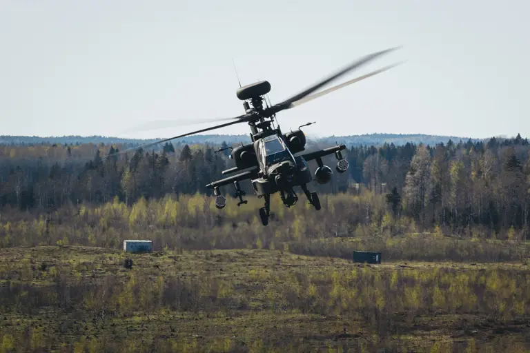 Military attack helicopter flying low over a grassy field with forest in the background on a clear day.