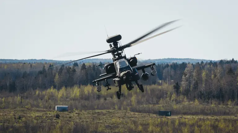 Military attack helicopter flying low over a grassy field with forest in the background on a clear day.
