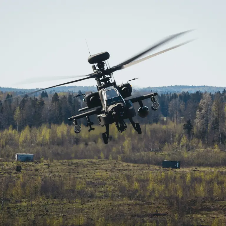 Military attack helicopter flying low over a grassy field with forest in the background on a clear day.
