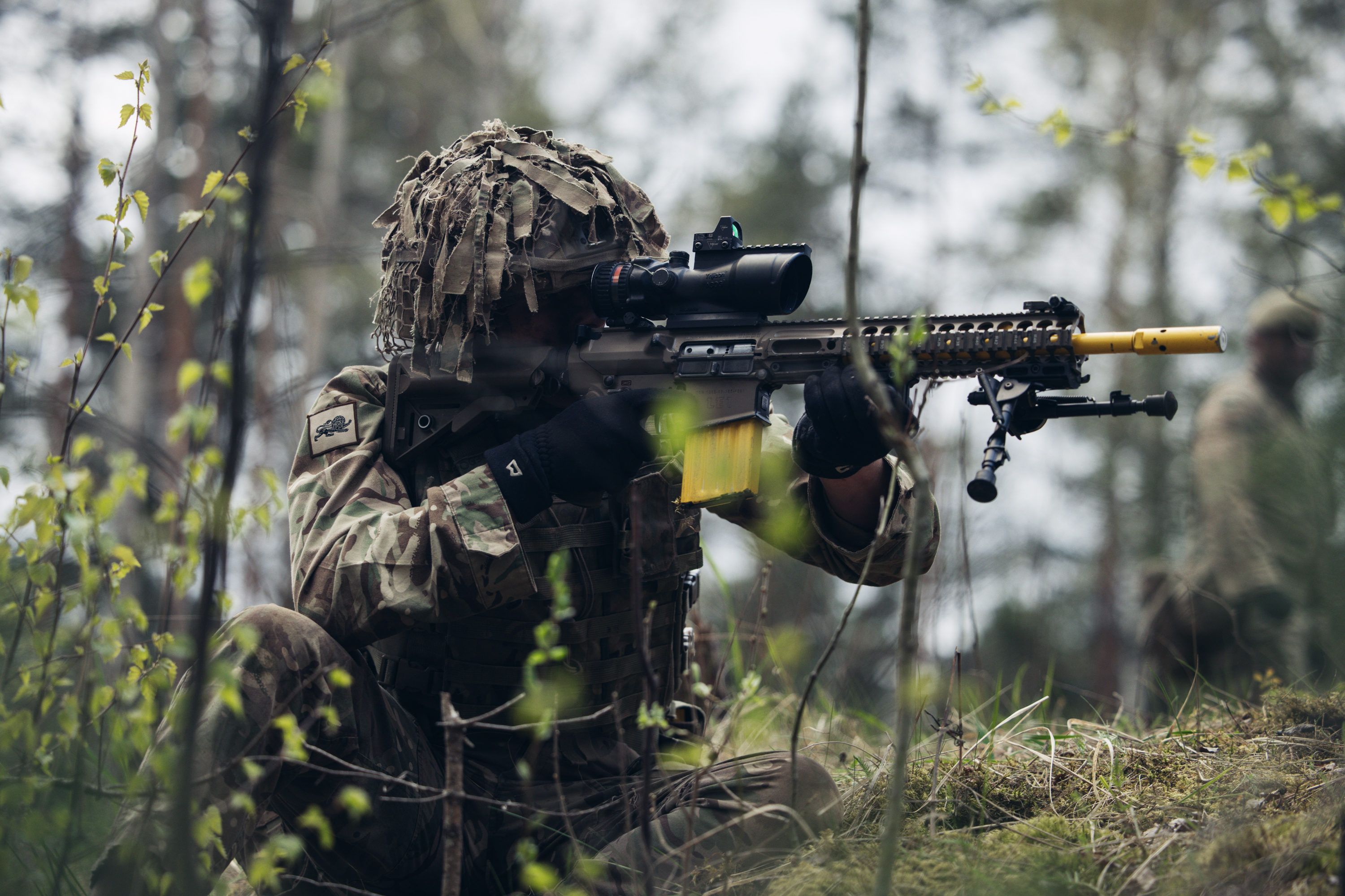 A soldier wearing camouflage uniform sits while aiming his rifle.