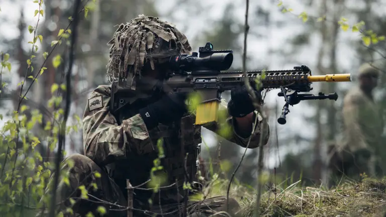 A soldier wearing camouflage uniform sits while aiming his rifle.