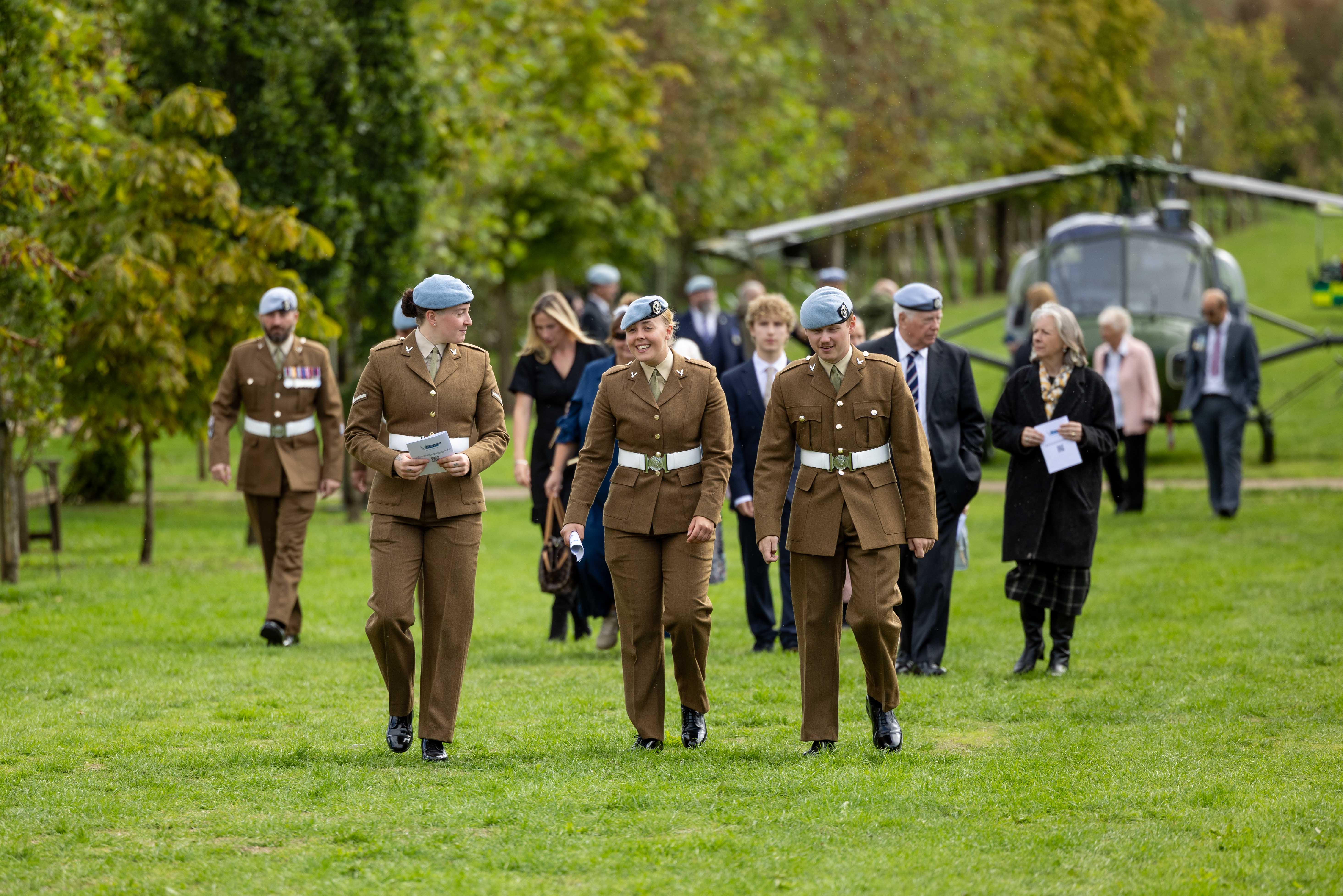 A group of people in military uniforms and formal attire walk across a grassy area. There is a helicopter in the background. Several individuals are wearing berets. The scene includes trees and a clear sky.