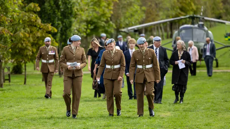 A group of people in military uniforms and formal attire walk across a grassy area. There is a helicopter in the background. Several individuals are wearing berets. The scene includes trees and a clear sky.
