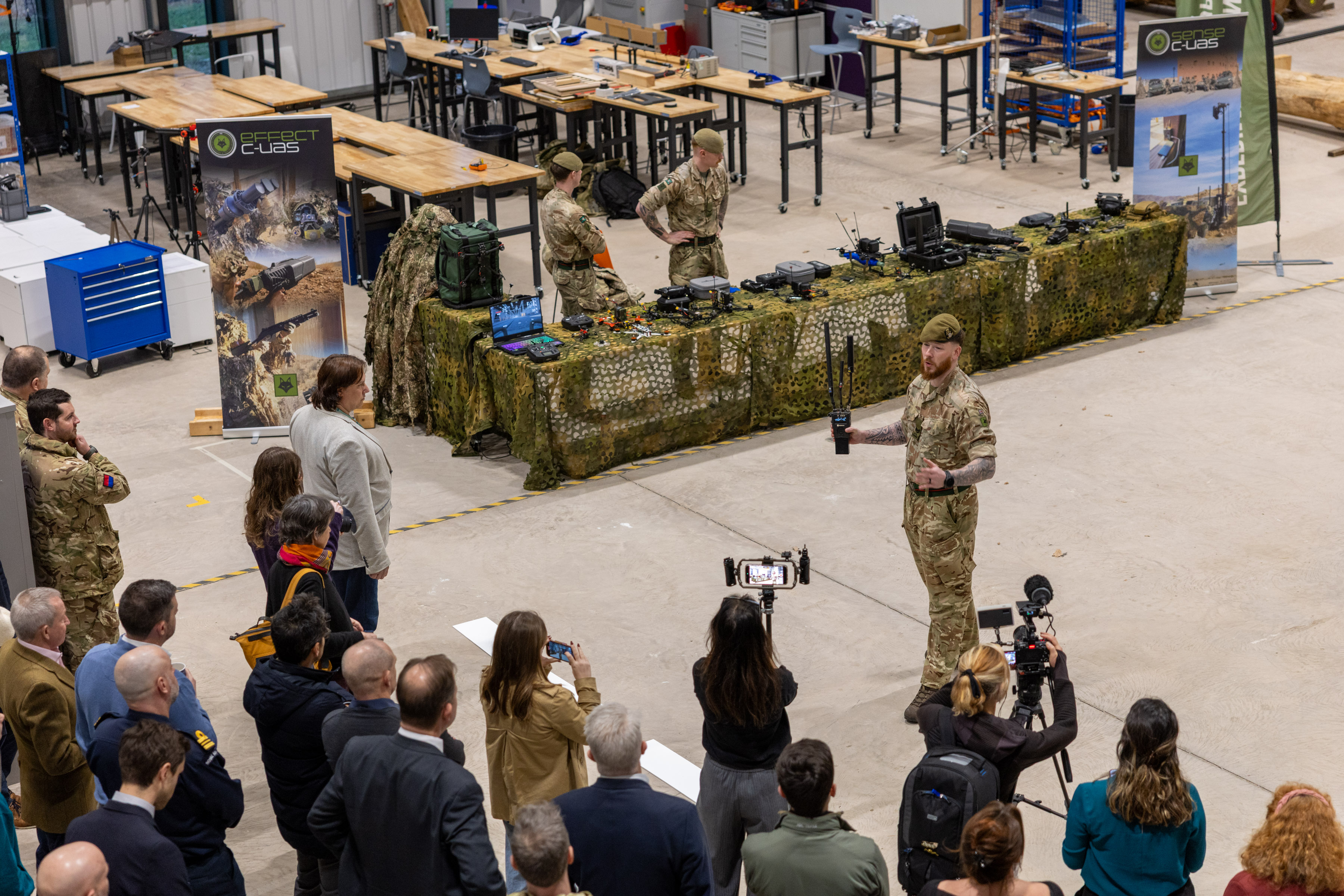 Military personnel demonstrating equipment to a crowd inside a large industrial space with camouflage-covered tables.