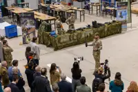 Military personnel demonstrating equipment to a crowd inside a large industrial space with camouflage-covered tables.