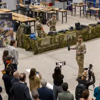 Military personnel demonstrating equipment to a crowd inside a large industrial space with camouflage-covered tables.