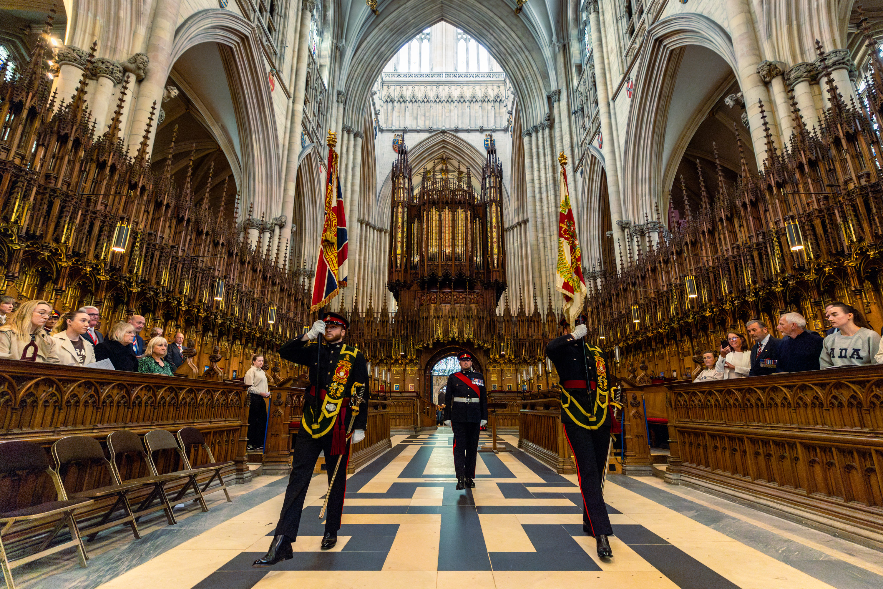 Ceremonial guards holding flags stand in the grand interior of York Minster. The cathedral's ornate architecture and tall arches are visible. Visitors are seated along the sides, observing the scene.
