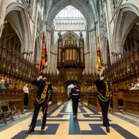 Ceremonial guards holding flags stand in the grand interior of York Minster. The cathedral's ornate architecture and tall arches are visible. Visitors are seated along the sides, observing the scene.
