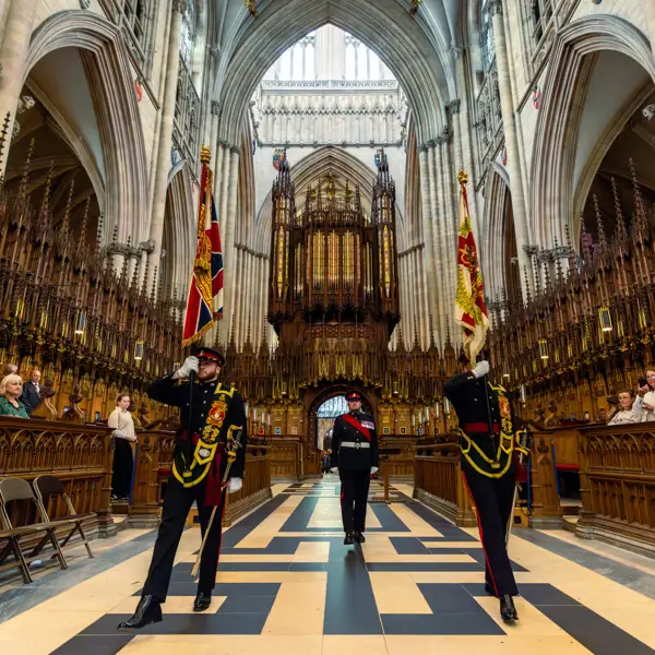 Ceremonial guards holding flags stand in the grand interior of York Minster. The cathedral's ornate architecture and tall arches are visible. Visitors are seated along the sides, observing the scene.