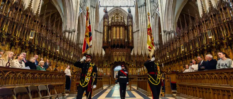 Ceremonial guards holding flags stand in the grand interior of York Minster. The cathedral's ornate architecture and tall arches are visible. Visitors are seated along the sides, observing the scene.