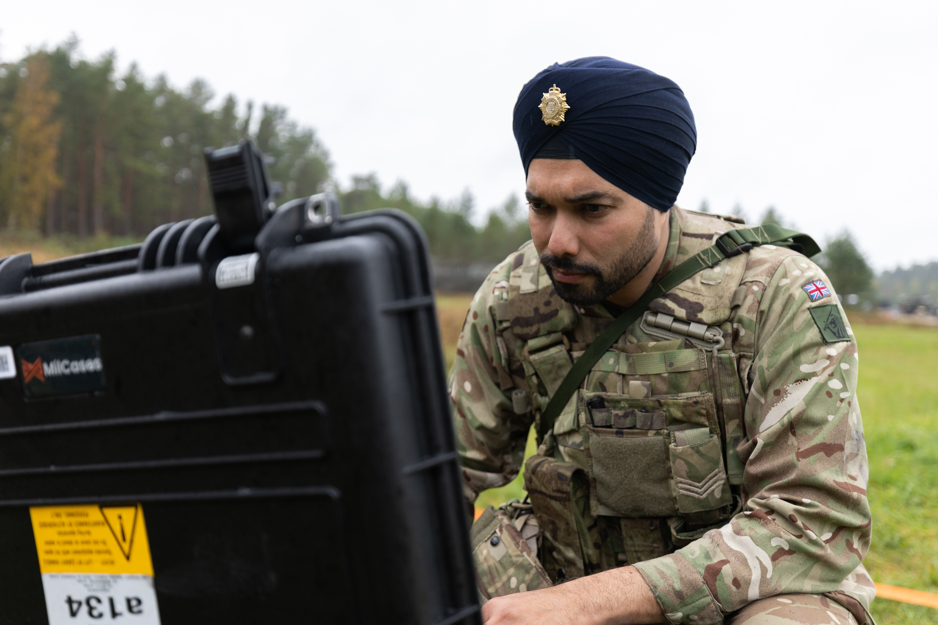 Soldier in camouflage uniform operating equipment outdoors in a grassy field with trees in the background.