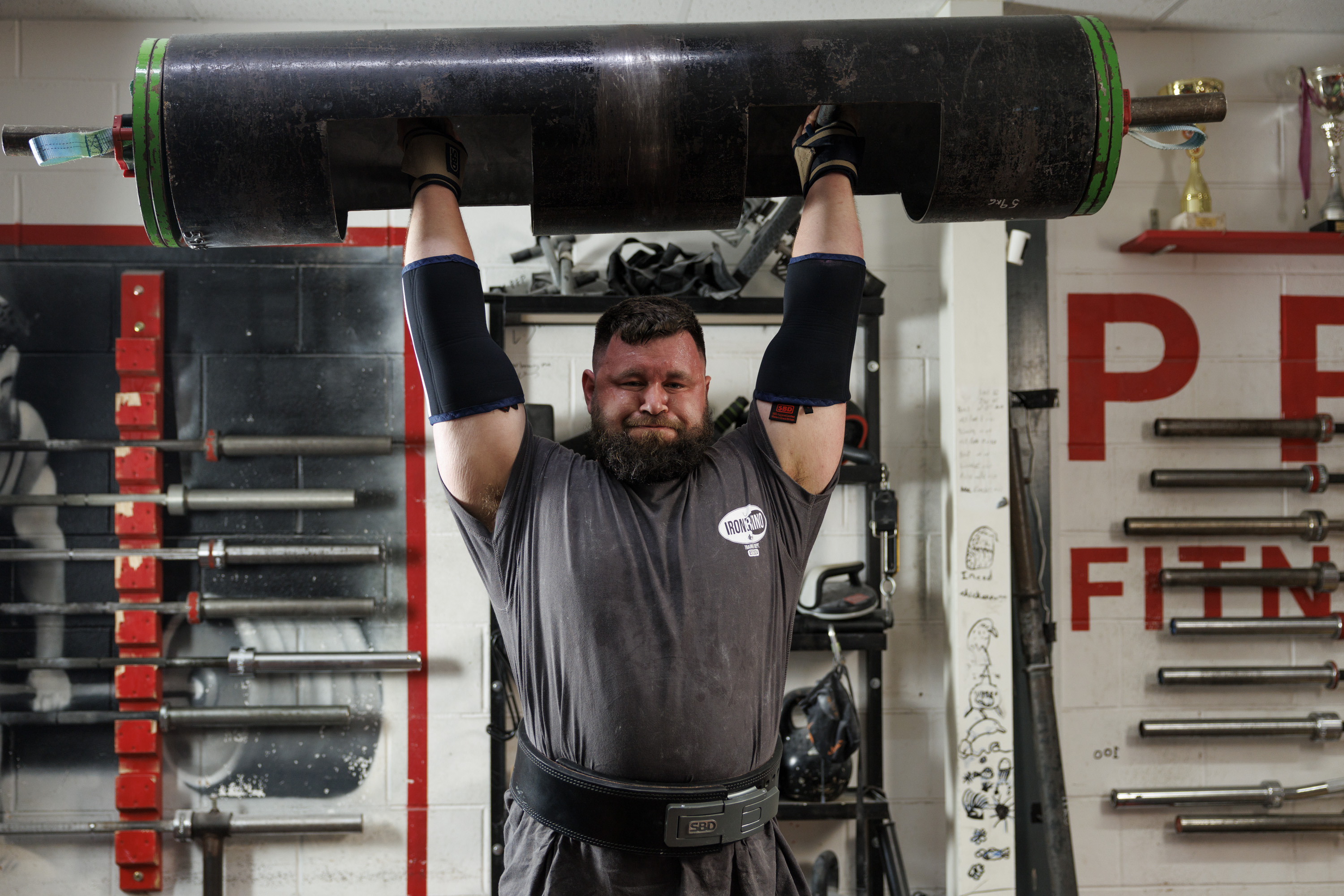 A person in a gym raises a large, heavy log overhead with both hands. The gym has various weightlifting equipment and a visible Power Fitness logo on the wall. The individual wears a dark shirt, protective sleeves, and a weightlifting belt.