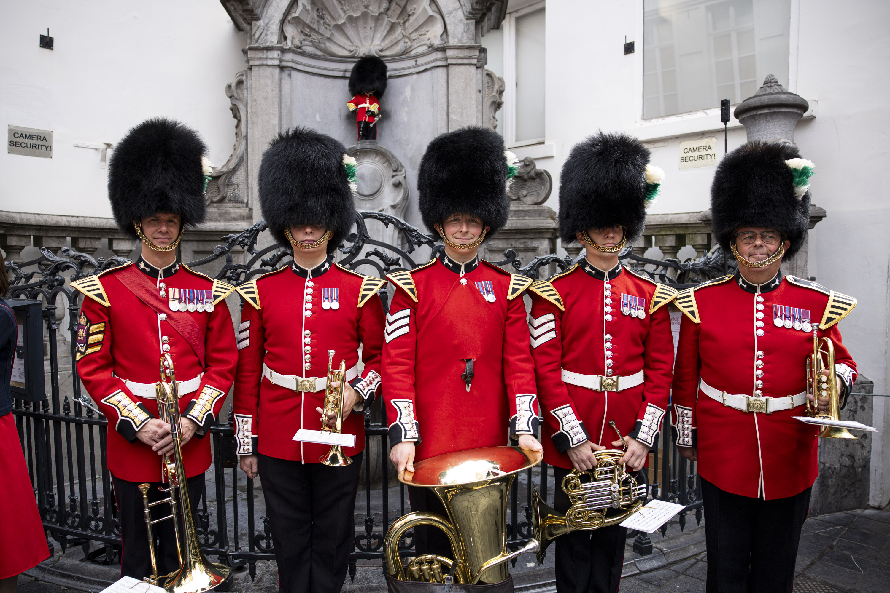 Five British guardsmen in traditional red uniforms and bearskin hats stand in front of a historic stone monument.