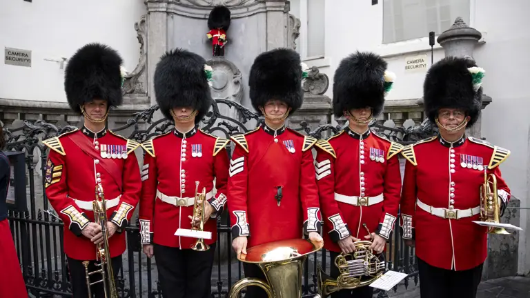 Five British guardsmen in traditional red uniforms and bearskin hats stand in front of a historic stone monument.