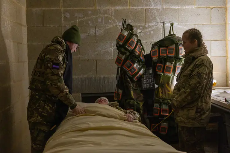 Two soldiers attend to a wounded comrade lying on a bed in a dimly lit concrete room.