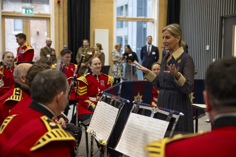 Her Royal Highness, The Duchess of Edinburgh, stands in front of a band of soldiers wearing red tunics and holding musical instruments.