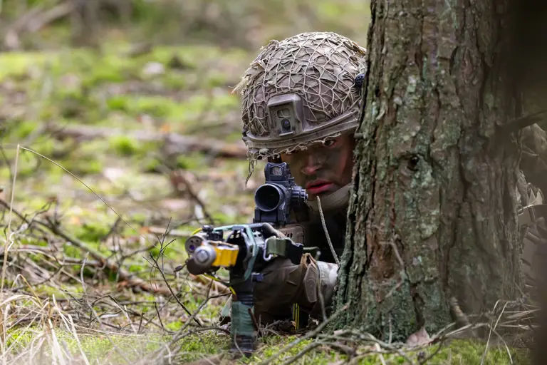 A soldier wearing a helmet takes aim with his rifle. He is laying down next to a tree.