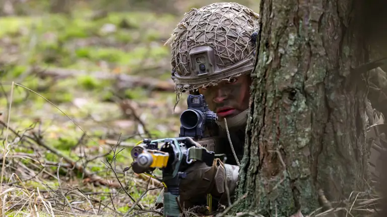 A soldier wearing a helmet takes aim with his rifle. He is laying down next to a tree.