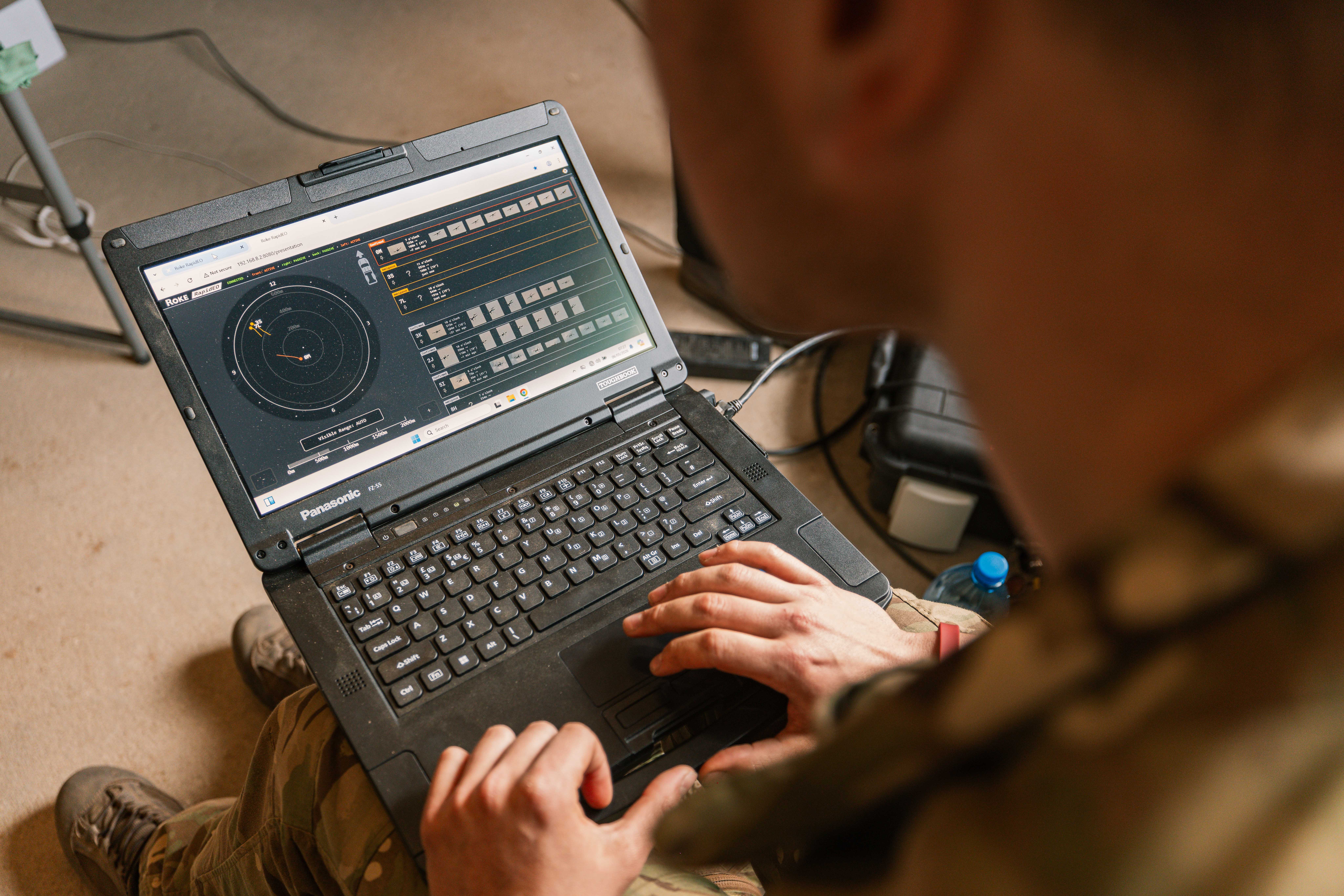 A person in military attire operates a rugged laptop displaying a radar interface, suggesting a tactical or strategic military setting.