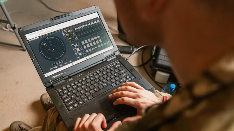A person in military attire operates a rugged laptop displaying a radar interface, suggesting a tactical or strategic military setting.