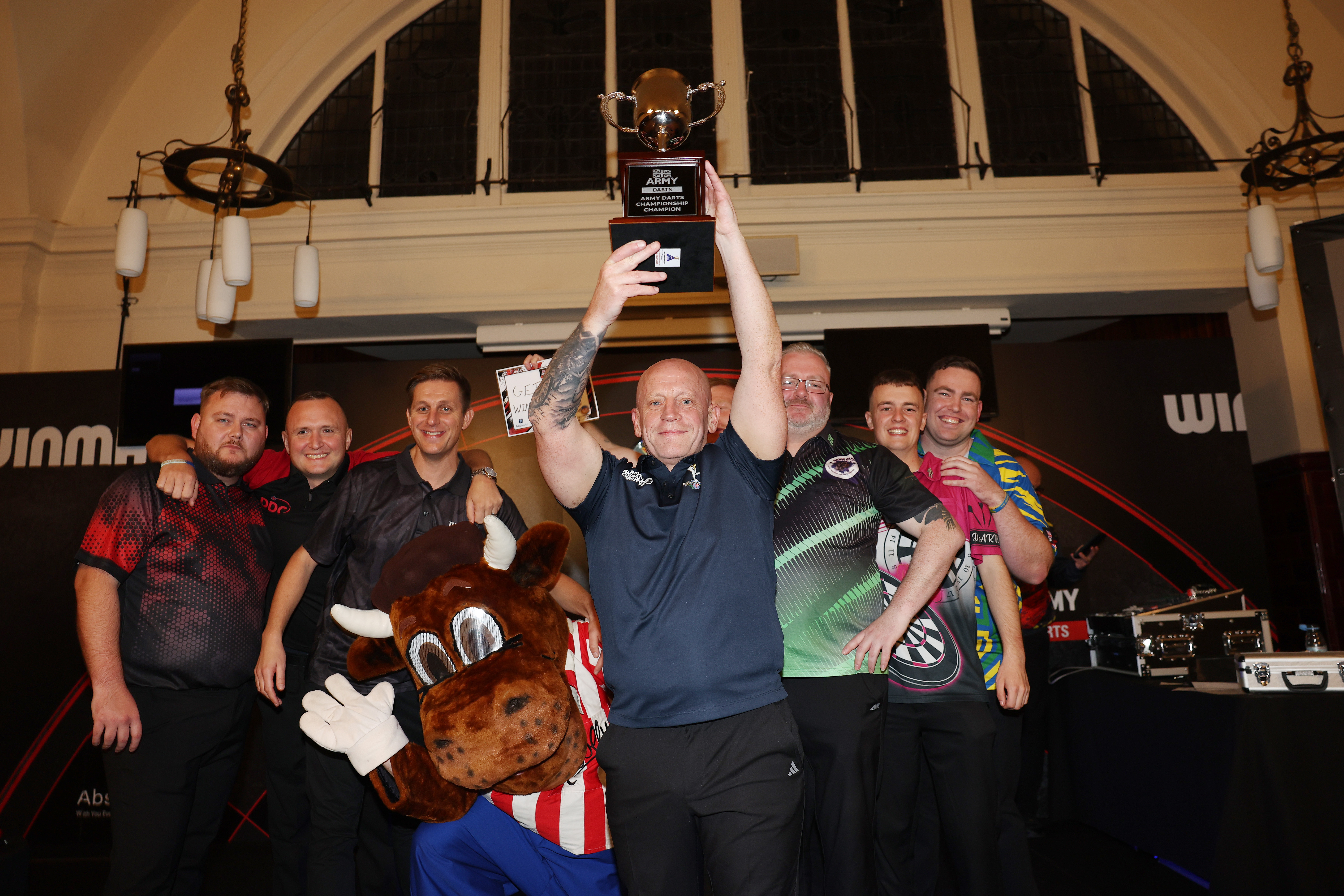 Group of six men and a mascot posing indoors, one man holding a trophy high above his head in celebration.
