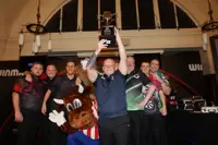 Group of six men and a mascot posing indoors, one man holding a trophy high above his head in celebration.