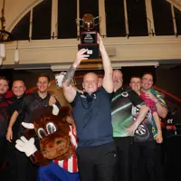 Group of six men and a mascot posing indoors, one man holding a trophy high above his head in celebration.