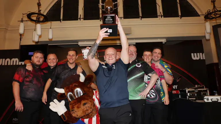 Group of six men and a mascot posing indoors, one man holding a trophy high above his head in celebration.