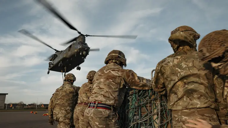 Soldiers wearing their camouflage uniform and helmets stand waiting with a load as a chinook coming into be loaded.