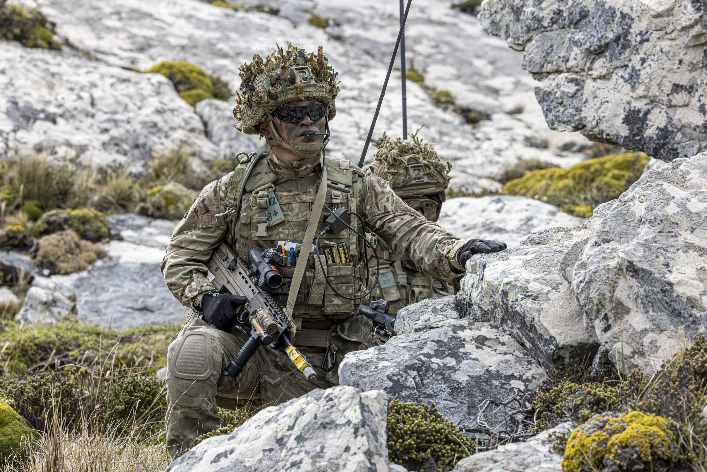 Two soldiers, wearing camouflage uniforms and gear, crouch in a rocky, moss-covered area. 