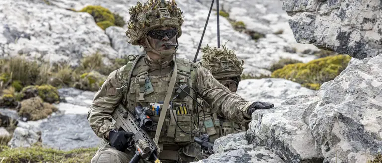 Two soldiers, wearing camouflage uniforms and gear, crouch in a rocky, moss-covered area.