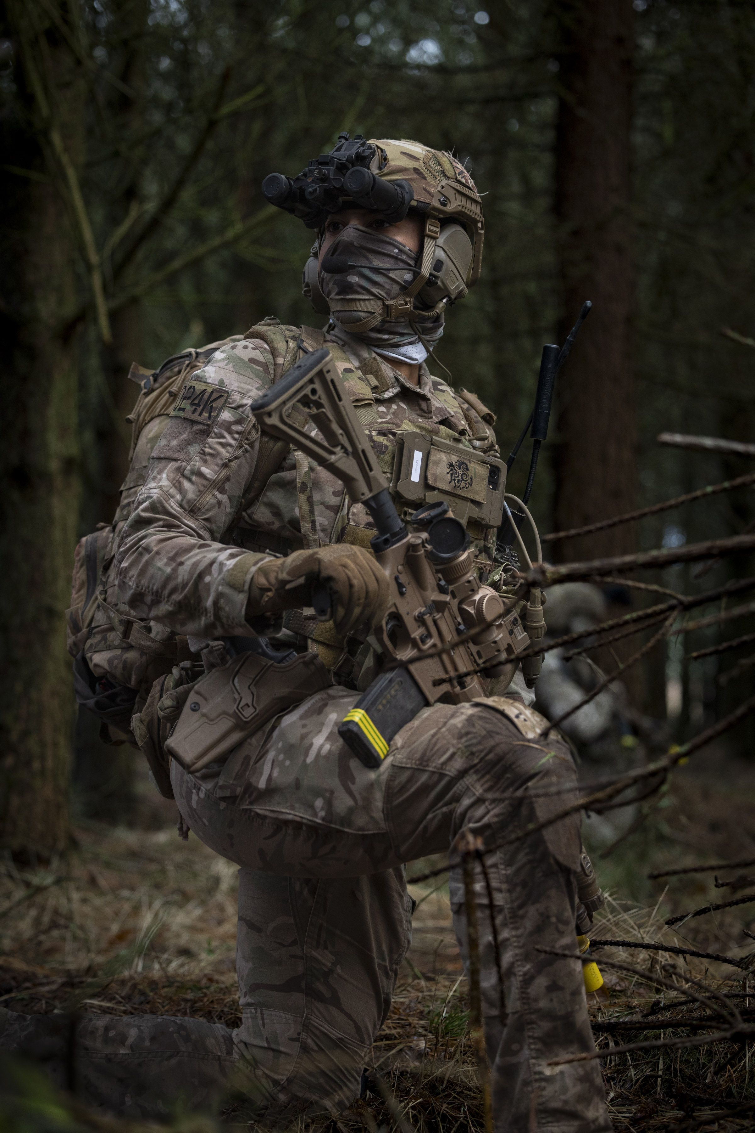 Soldier in full tactical gear kneeling in a forest, holding a rifle with advanced attachments and wearing camouflage.