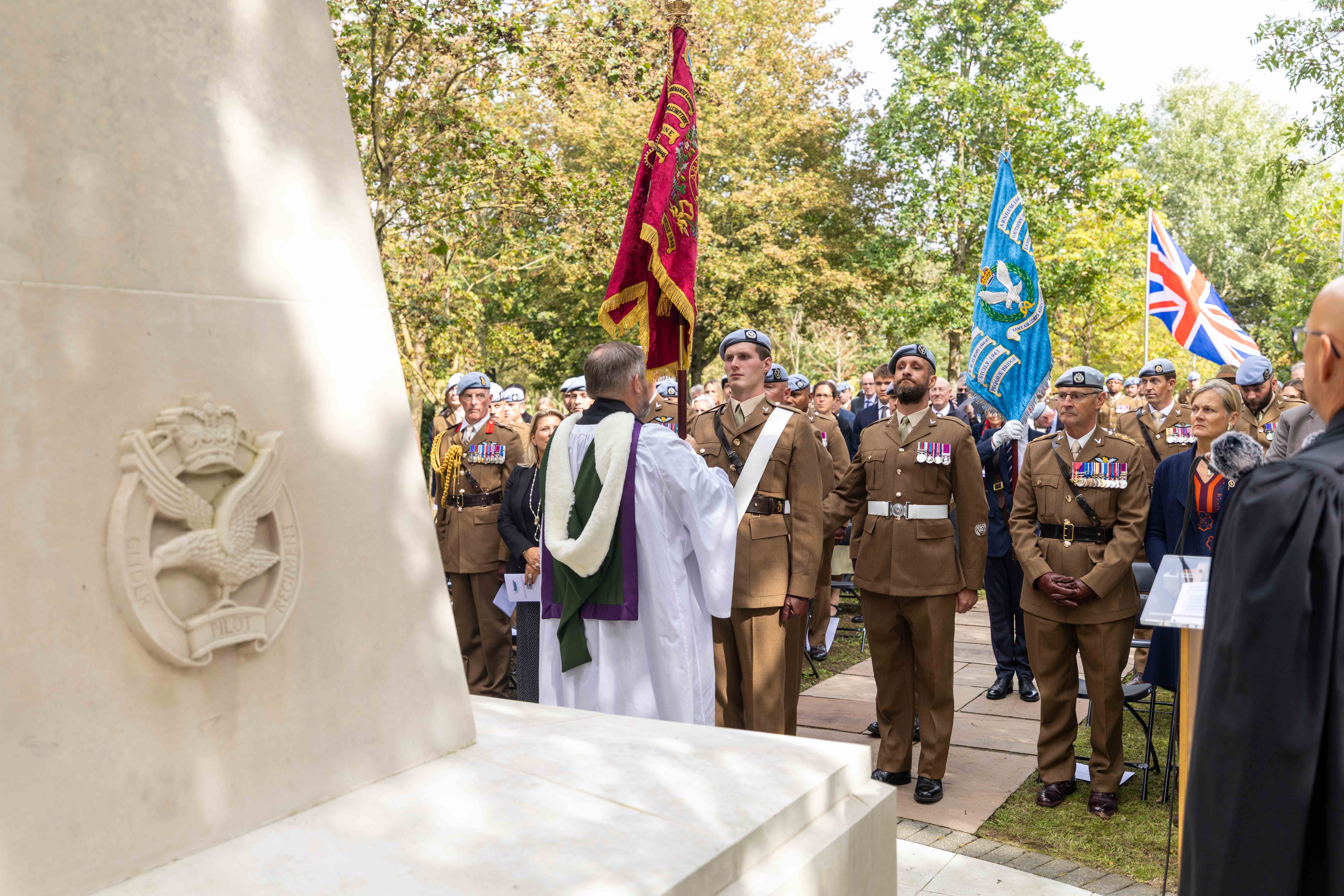 A ceremonial gathering by a large memorial, featuring people in military uniforms holding flags, including the Union Jack. Trees provide a backdrop. A person in robes stands near the memorial, addressing the group.