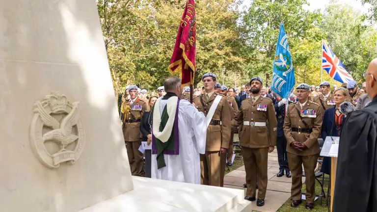 A ceremonial gathering by a large memorial, featuring people in military uniforms holding flags, including the Union Jack. Trees provide a backdrop. A person in robes stands near the memorial, addressing the group.