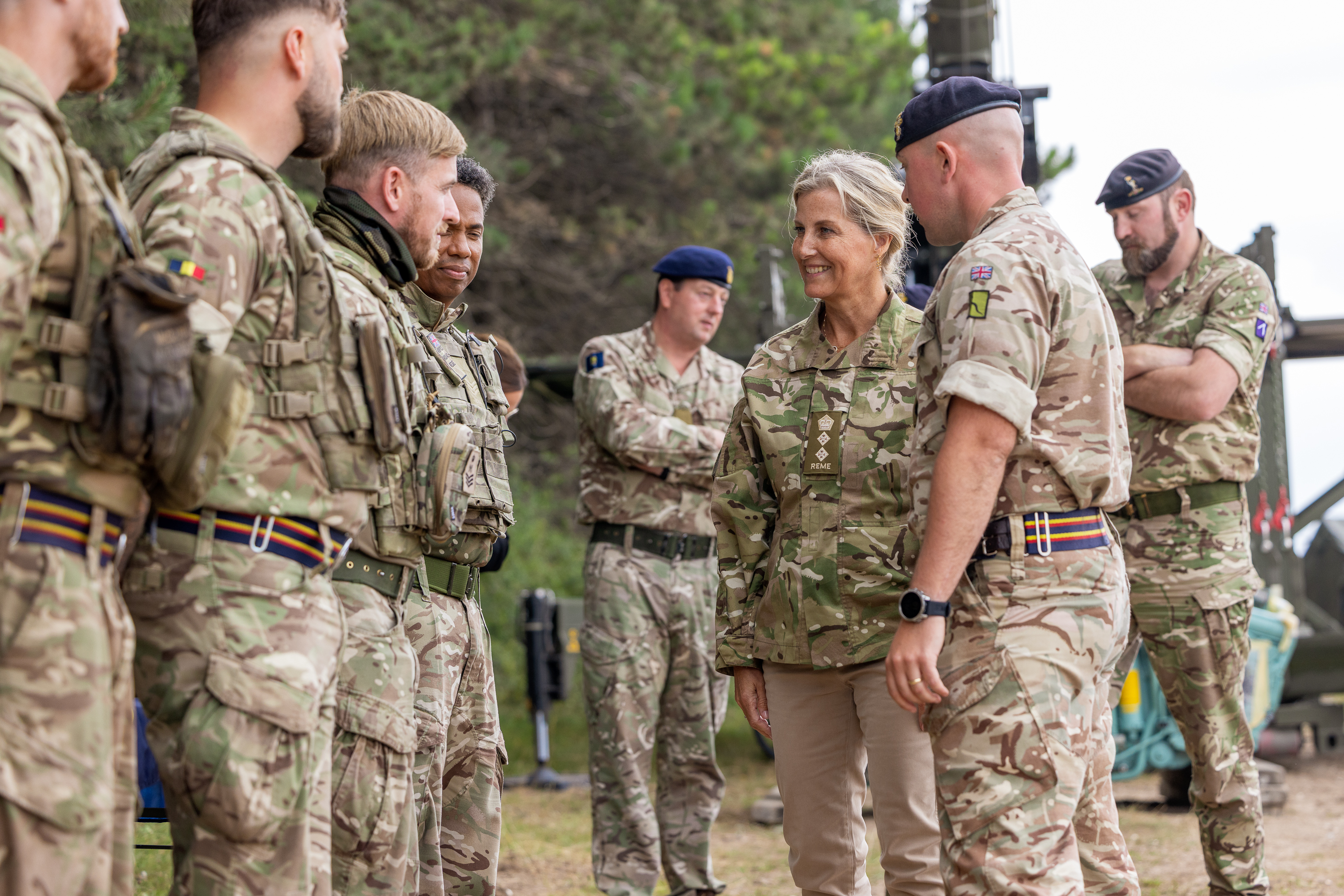 The Duchess of Edinburgh is seen wearing trousers and a camouflage print jacket stood walking outside with two soldiers in camouflage uniform on either side of her. smiling at a line of soldiers in front of her