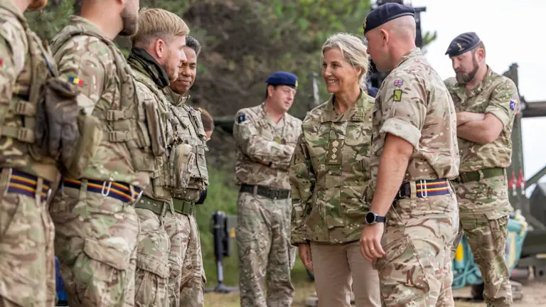 The Duchess of Edinburgh is seen wearing trousers and a camouflage print jacket stood walking outside with two soldiers in camouflage uniform on either side of her. smiling at a line of soldiers in front of her