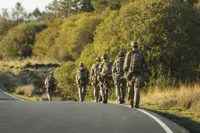 A group of soldiers in camouflage walks along a winding road amid lush, sunlit greenery.
