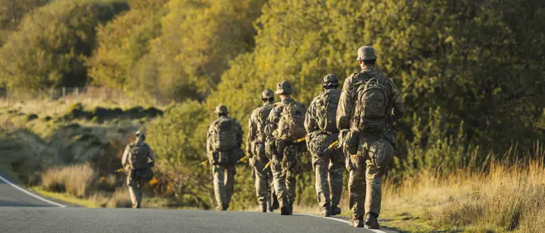 A group of soldiers in camouflage walks along a winding road amid lush, sunlit greenery.