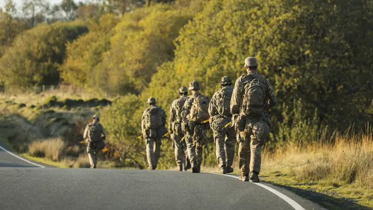 A group of soldiers in camouflage walks along a winding road amid lush, sunlit greenery.