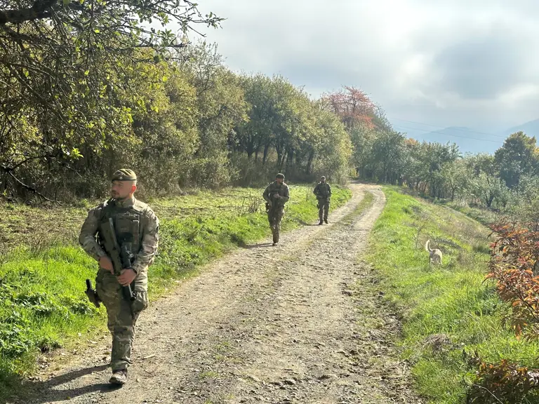 Three soldiers wearing camouflage uniforms and carrying rifles walk along a gravel track.