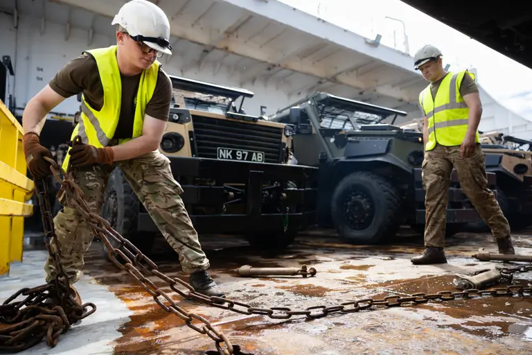 A soldier wearing a hi vis moves a chain aboard a ferry. Behind him are a row of green and beige military vehicles.