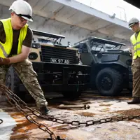 A soldier wearing a hi vis moves a chain aboard a ferry. Behind him are a row of green and beige military vehicles.