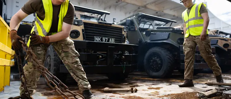 A soldier wearing a hi vis moves a chain aboard a ferry. Behind him are a row of green and beige military vehicles.
