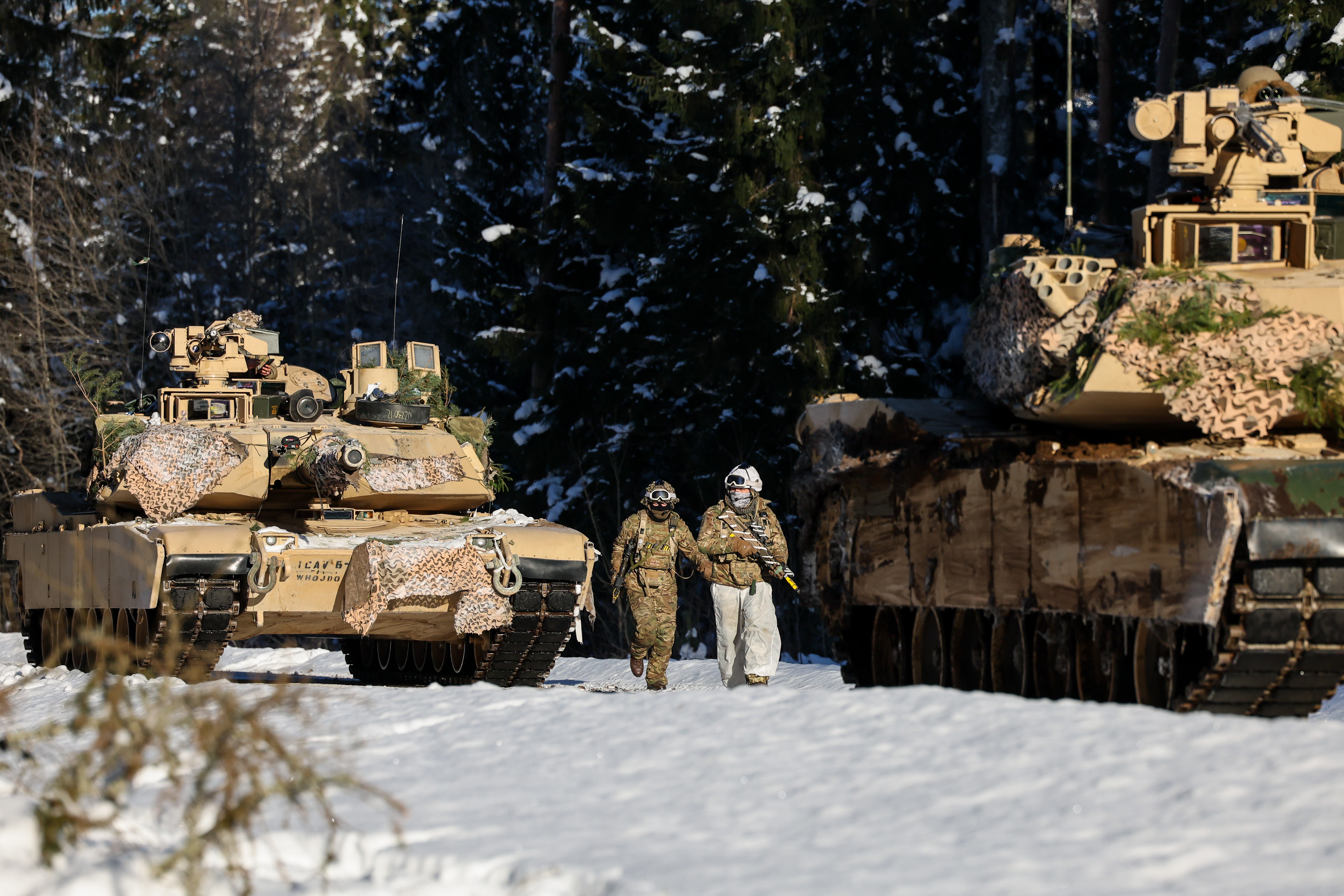 Two soldiers in winter camouflage walking between camouflaged military tanks in a snowy forest clearing.