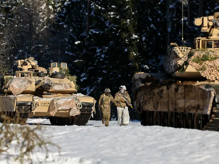 Two soldiers in winter camouflage walking between camouflaged military tanks in a snowy forest clearing.
