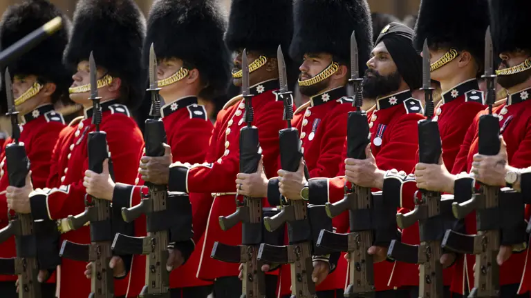 A line of soldiers in red tunics and bearskin hats hold their rifles towards the sky.
