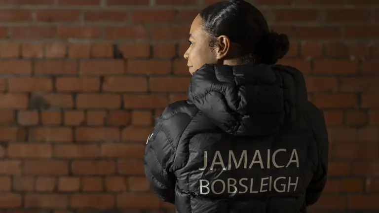 Person wearing a black puffer jacket with 'Jamaica Bobsleigh' printed on the back, standing against a brick wall.