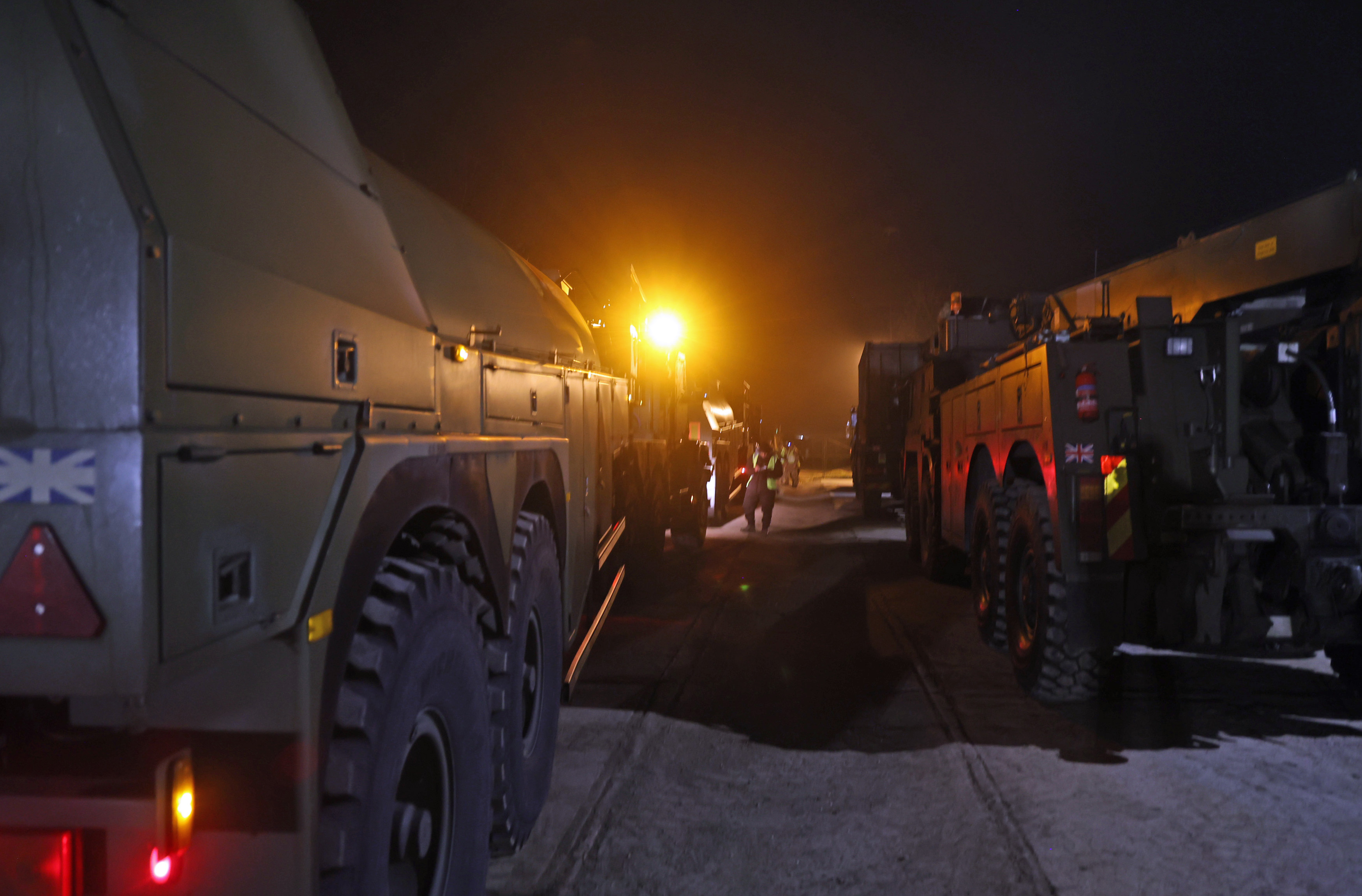 Two rows of large army transport vehicles parked up waiting for transport, inspected by a soldier in camouflage uniform. 