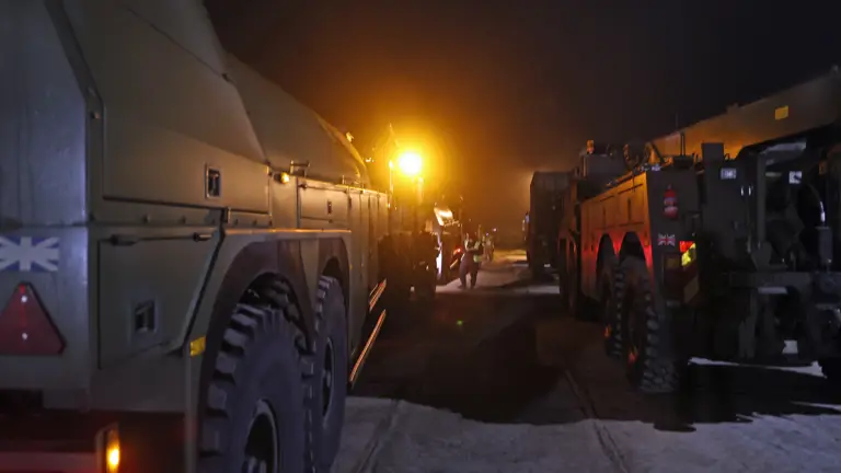 Two rows of large army transport vehicles parked up waiting for transport, inspected by a soldier in camouflage uniform.