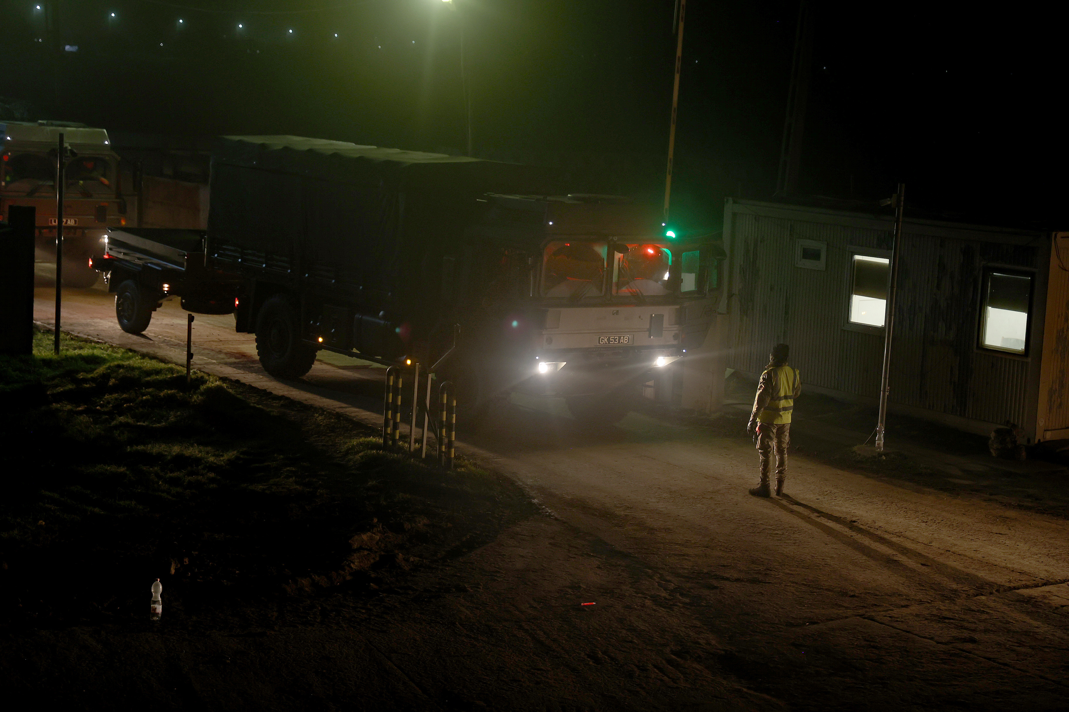 Large army transport vehicle being guided by a soldier in camouflage uniform at night.